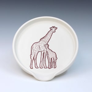 May include: White ceramic spoon rest with a brown giraffe illustration. The design features a mother giraffe and a baby giraffe. The spoon rest has a curved shape with a small lip for resting utensils.