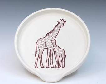 May include: White ceramic spoon rest with a brown giraffe illustration. The design features a mother giraffe and a baby giraffe. The spoon rest has a curved shape with a small lip for resting utensils.