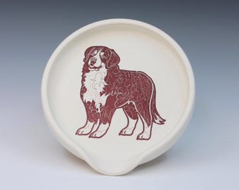 May include: A white ceramic spoon rest with a maroon dog illustration. The dog is standing and has detailed patterns within its fur. The spoon rest has a rounded shape with a small spout for resting utensils.