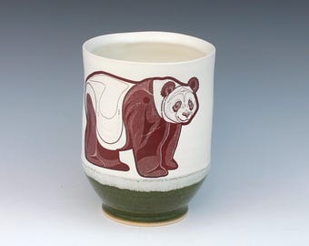 May include: A white ceramic mug with a hand-painted panda bear design. The panda is brown and white, with a detailed face. The bottom of the mug is green, and the interior is white.