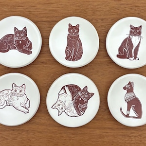 May include: Six white ceramic coasters with brown cat illustrations. Each coaster features a different cat design, including cats sitting, lying down, and with decorative patterns. The coasters are arranged on a wooden surface.