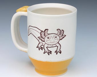 May include: A white ceramic mug featuring a brown line drawing of an axolotl on the front. The base of the mug is glazed in a solid yellow color, and the handle is attached to a yellow decorative accent at the top. The interior is white.