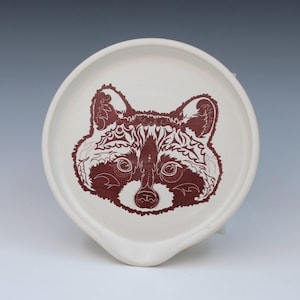 May include: A round, off-white ceramic spoon rest featuring a detailed, dark reddish-brown illustration of a raccoon face in the center. The raccoon design incorporates intricate, stylized patterns within its fur and facial features on the smooth surface.
