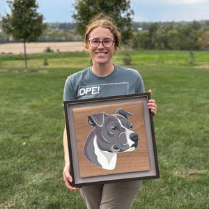 May include: A framed artwork featuring a stylized portrait of a dog's head, rendered in shades of gray, white, and purple against a wood-grain background. The artwork is encased in a black frame with a gray matting.