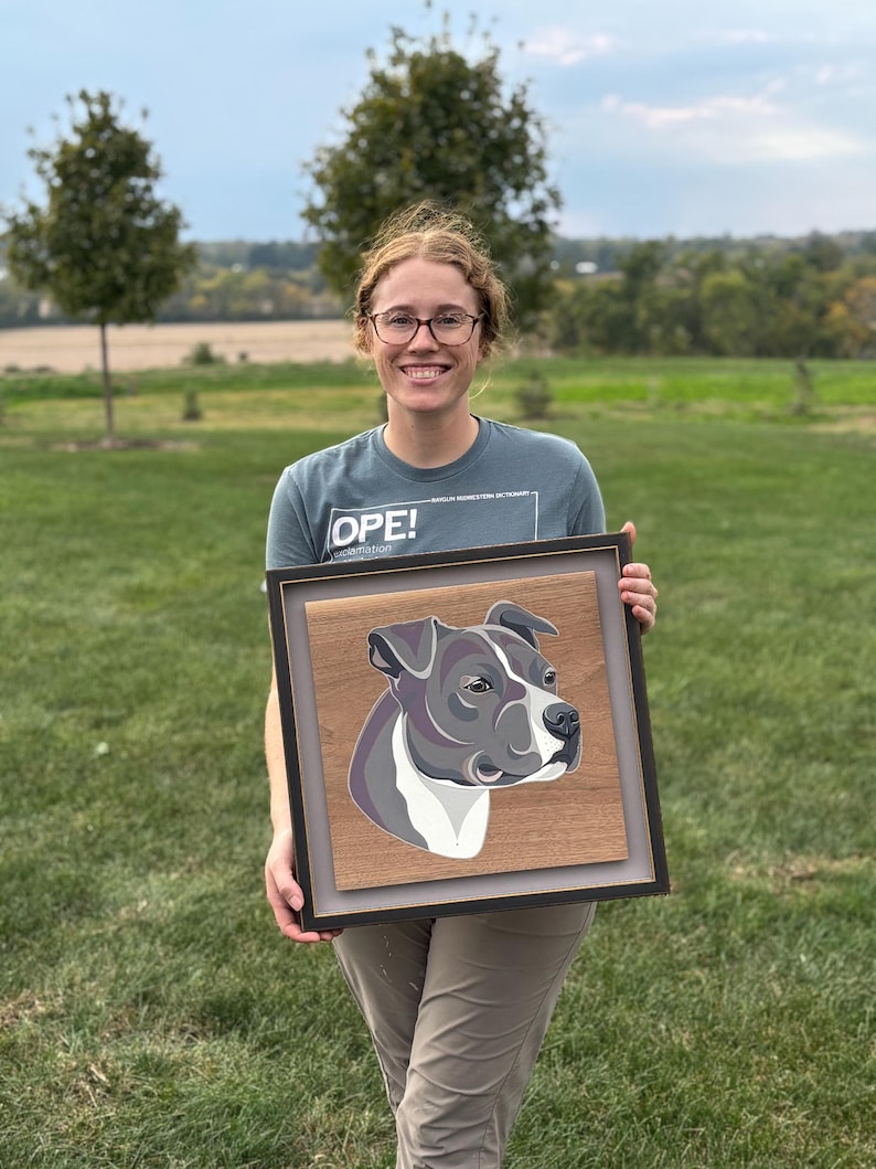 May include: A framed artwork featuring a stylized portrait of a dog's head, rendered in shades of gray, white, and purple against a wood-grain background. The artwork is encased in a black frame with a gray matting.