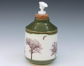 May include: A ceramic soap dispenser with a white cylindrical body featuring dark red tree silhouettes and a crescent moon. The top and base are glazed in a deep forest green. A cork stopper sits at the neck, topped with a white plastic pump.