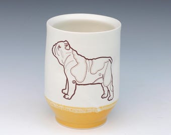 May include: A white ceramic tumbler with a brown outline drawing of a bulldog. The bottom of the tumbler is a light yellow color. The tumbler is cylindrical and has a smooth surface.