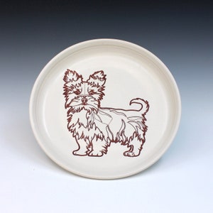 May include: A cream-colored ceramic plate featuring a brown outline illustration of a Yorkshire Terrier dog. The dog is standing, with detailed fur and a small tail. The plate has a simple, rounded rim.