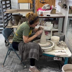 May include: A person working on a pottery wheel, shaping a clay vase. The studio setting includes shelves with pottery, tools, and a folding chair. The person is wearing headphones and a green shirt.
