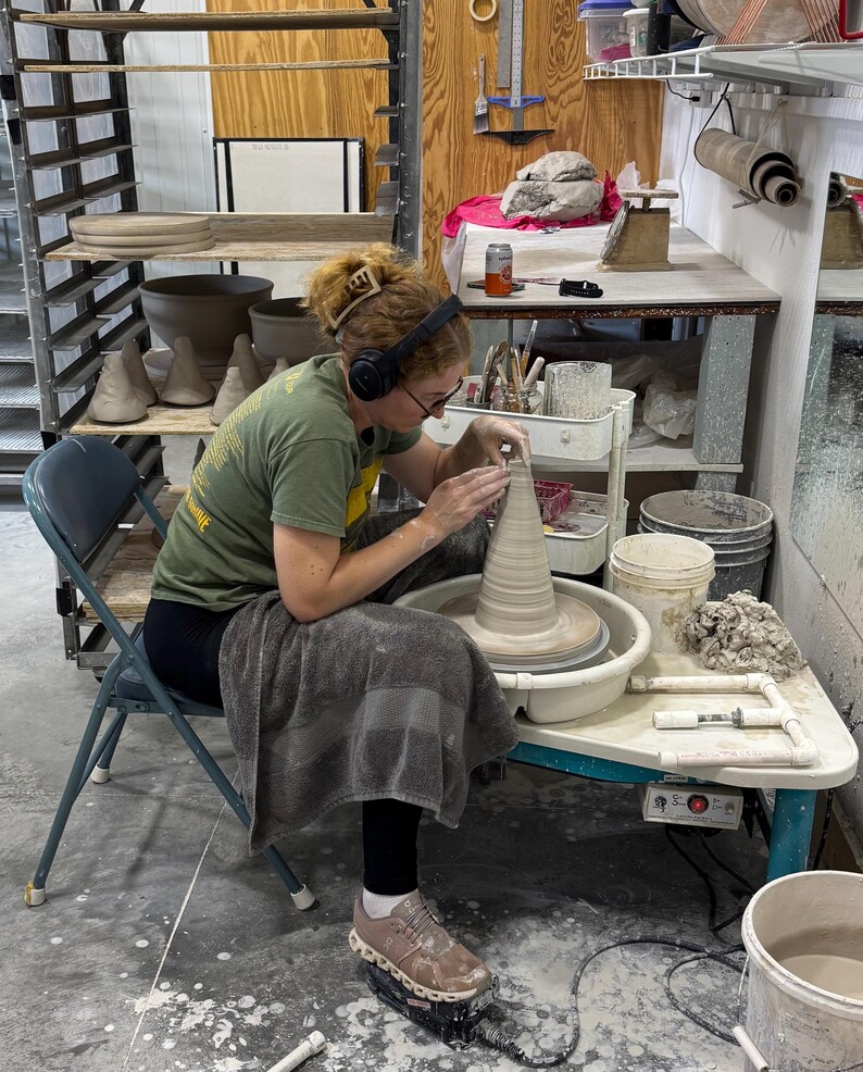 May include: A person working on a pottery wheel, shaping a clay vase. The studio setting includes shelves with pottery, tools, and a folding chair. The person is wearing headphones and a green shirt.