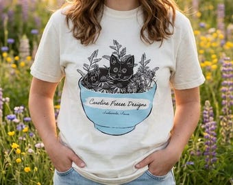May include: A cream-colored short-sleeve t-shirt featuring a graphic of a black cat sitting inside a light blue bowl filled with flowers. The bowl displays the text Caroline Freen Designs and Asheville, North Carolina in black script.