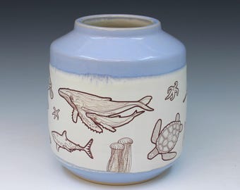 May include: A ceramic vase with a light blue glazed top and bottom and a white center band. The white section features brown line drawings of marine life, including a whale, a sea turtle, a shark, starfish, and coral structures on a smooth surface.