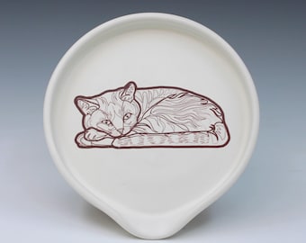 May include: A round, off-white ceramic spoon rest featuring a dark brown line art illustration of a cat lying down. The cat is depicted in a resting position with its head tucked near its front paws, showcasing detailed fur texture and markings.