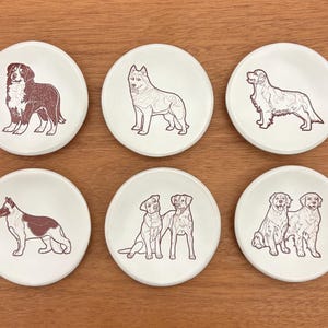 May include: Set of six white ceramic coasters featuring dog illustrations in brown. Each coaster showcases a different breed, including a Bernese Mountain Dog, Husky, Golden Retriever, German Shepherd, and two Labrador Retrievers.