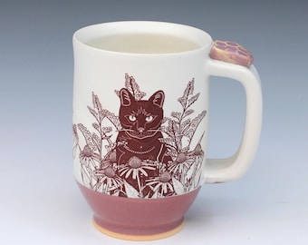 May include: A white ceramic mug featuring a dark brown line art illustration of a cat sitting among flowers. The base of the mug is a muted pink color, and the handle is topped with a small, textured, pink and yellow honeycomb-shaped accent.