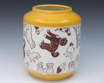 May include: A cylindrical ceramic vase with a bright yellow glazed rim and base. The central white body features a repeating pattern of various dog breeds illustrated in dark brown ink, showcasing different poses and orientations across the surface.