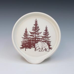 May include: A white ceramic spoon rest with a brown illustration of three pine trees and a bear. The spoon rest has a curved shape with a small lip for resting utensils. The design is rustic and nature-themed.