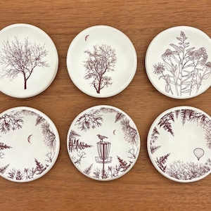 May include: Set of six white ceramic coasters with nature-themed illustrations in dark red. Designs include trees, flowers, a bird on a disc golf basket, and a hot air balloon. Coasters are round and sit on a wooden surface.