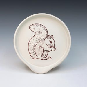 May include: Off-white ceramic spoon rest with a brown squirrel illustration. The squirrel is depicted in a detailed, stylized design, with a large, bushy tail and intricate patterns. The spoon rest has a curved shape with a small lip.