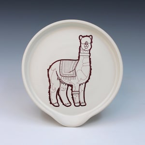 May include: A white ceramic spoon rest with a brown outline drawing of an alpaca. The alpaca is wearing a saddle and has decorative markings. The spoon rest has a curved shape with a small lip for resting utensils.