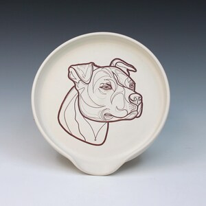 May include: Cream-colored ceramic spoon rest with a brown line drawing of a dog's head. The spoon rest has a rounded shape with a small indentation for resting utensils.