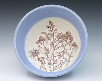 May include: A round ceramic bowl with a light blue exterior and interior rim. The center of the bowl features a detailed brown line drawing of various wildflowers and foliage on a cream-colored background, showcasing botanical stems and petals.