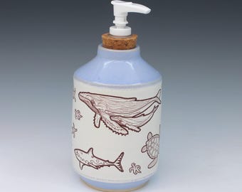 May include: A ceramic soap dispenser with a light blue top and base, featuring a white center band decorated with brown line drawings of a whale, a sea turtle, a shark, and starfish. The dispenser has a cork stopper and a white plastic pump.