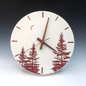 May include: A round, cream-colored clock with a forest scene. The clock face features a crescent moon and dark red silhouettes of pine trees. The clock hands and hour markers are brown. The clock is a decorative item.
