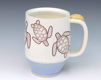 May include: A ceramic mug with a white upper body featuring brown line art of sea turtles. The lower portion of the mug is light blue, and the handle is topped with a small, textured yellow accent piece. The interior of the mug is white.