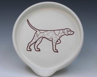 May include: A round, off-white ceramic spoon rest featuring a dark brown line drawing of a dog in profile. The dog illustration is filled with a pattern of small circles and connected lines, resembling a constellation map on the animal's body.