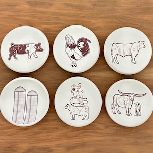 May include: Set of six ceramic coasters with farm animal illustrations in brown on a cream background. Designs include a pig, rooster, bull, silos, a stack of animals, and a longhorn with a calf. Coasters are round.
