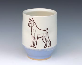 May include: A white ceramic tumbler with a light blue base and a brown outline drawing of a Boxer dog. The dog illustration is centered on the tumbler, with a spiral design on its hindquarters.