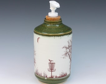 May include: A ceramic soap dispenser with a white pump. The cylindrical body is white with a green top and base. It features a red illustration of a disc golf basket, trees, and a bird. The dispenser is approximately 7 inches tall.