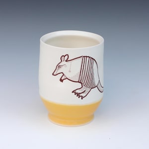 May include: A ceramic tumbler with a white upper section and a yellow base. A brown illustration of an armadillo is on the white section. The tumbler has a cylindrical shape and a slightly flared base.