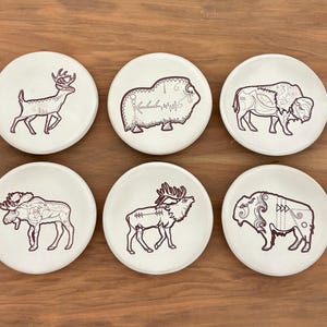 May include: Six white ceramic coasters with brown animal illustrations. The coasters feature a deer, musk ox, and bison, along with a moose and two other bison. The coasters are round and sit on a wooden surface.
