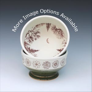 May include: A ceramic bowl with a white interior featuring a brown forest and crescent moon illustration. The exterior has a white upper band with brown flower patterns and a dark green base. The text "More Image Options Available" is visible.