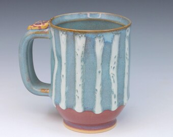 May include: A ceramic mug featuring a light blue glaze with vertical white drip patterns. The base of the mug is a solid reddish-brown color. A sturdy handle is attached to the side, topped with a small, textured, yellow and purple decorative accent.