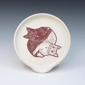 May include: A white ceramic spoon rest with a circular design featuring two cats in a yin-yang style. The cats are in a reddish-brown color with intricate patterns. The spoon rest has a curved handle.