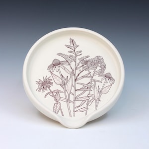 May include: Off-white ceramic spoon rest with a floral design in dark brown. The design features detailed illustrations of various wildflowers and foliage. The spoon rest has a curved shape with a small lip for holding utensils.