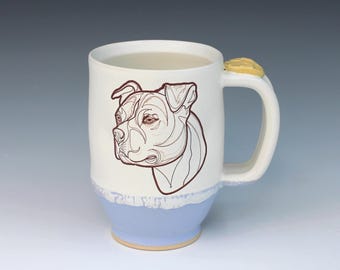 May include: A white ceramic mug featuring a brown line art illustration of a pit bull dog head on the front. The base of the mug is glazed in a light blue color with a textured drip effect, and the handle includes a small yellow accent piece.