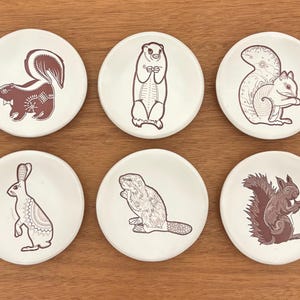 May include: Six white ceramic coasters with brown illustrations of woodland animals: a skunk, groundhog, squirrel, rabbit, beaver, and another squirrel. The coasters are arranged on a wooden surface.