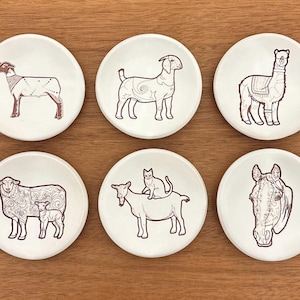 May include: Set of six white ceramic coasters with brown line drawings of farm animals. The designs include a sheep, goat, llama, horse, and a goat with a cat on its back. The coasters are round and sit on a wooden surface.