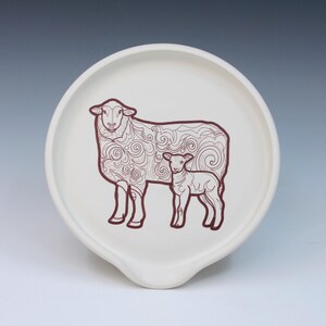 May include: A white ceramic spoon rest with a dark red illustration of a sheep and a lamb. The sheep has decorative swirls and lines. The spoon rest has a curved indentation for holding utensils.