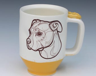 May include: A white ceramic mug featuring a dark brown line art illustration of a pit bull dog head on the front. The base of the mug is glazed in a solid yellow color, and the handle includes a small, raised yellow paw print detail at the top.