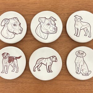 May include: Set of six white ceramic coasters featuring line drawings of different dog breeds in brown. The dogs are depicted in various poses, including standing and sitting. The coasters are arranged on a wooden surface.