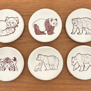 May include: Set of six white ceramic coasters featuring animal illustrations. Designs include a red panda, panda bear, bear with cub, tiger, bear with cub, and bear. Each coaster has a brown outline.
