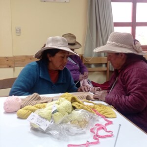 Pu&ograve; includere: Tre persone sono sedute a un tavolo a lavorare a maglia con filati di vari colori, tra cui rosa, giallo e marrone. Stanno usando ferri da maglia e un metro a nastro.