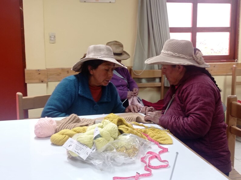 Pu&ograve; includere: Tre persone sono sedute a un tavolo a lavorare a maglia con filati di vari colori, tra cui rosa, giallo e marrone. Stanno usando ferri da maglia e un metro a nastro.
