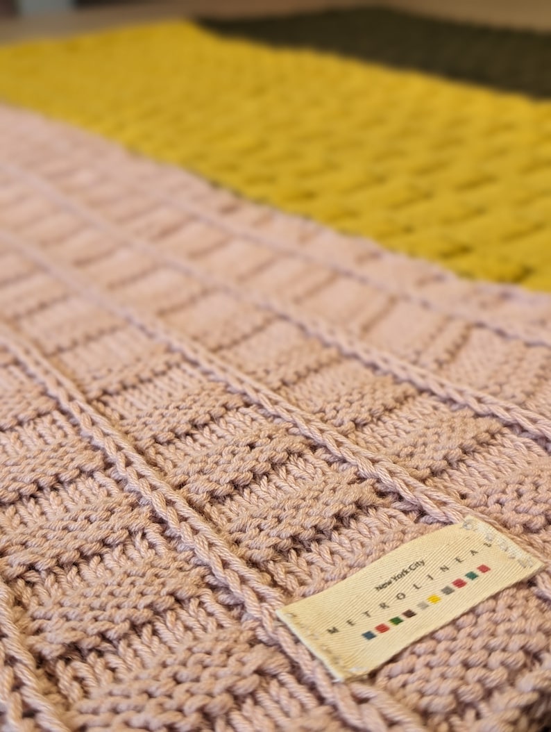 Pu&ograve; includere: Un primo piano di una sciarpa di maglia rosa con un'etichetta bianca che recita "New York City METRO LINEN". L'etichetta presenta un design a righe colorate.