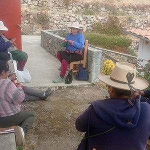 Pu&ograve; includere: Cinque persone sono sedute all'aperto, indossando cappelli e lavorando a maglia. Sono sedute su un patio con un muro di pietra e un tetto di tegole sullo sfondo.